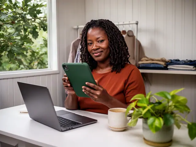 A boutique owner at a white desk holding a tablet in a green case, with a laptop, a mug and a pothos plant, clothes on a rack behind her.